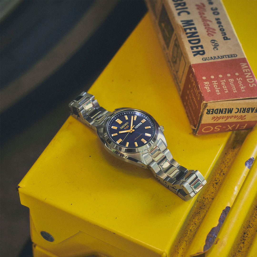 Seiko 5 Sports SRE021K1 navy blue dial watch in flatlay on a yellow toolbox, styled with vintage Fabric Mender packaging
