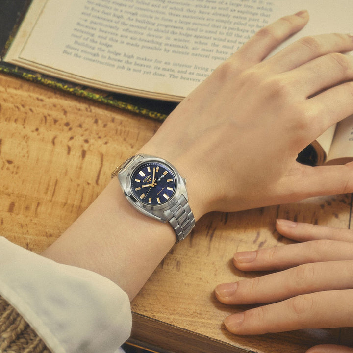 Seiko 5 Sports SRE021K1 navy dial watch on wrist, resting on an open vintage book, paired with a white long-sleeve top