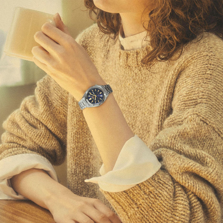 Seiko 5 Sports SRE021K1 navy dial watch worn on wrist, styled with a chunky camel knit sweater and white shirt, holding a warm drink