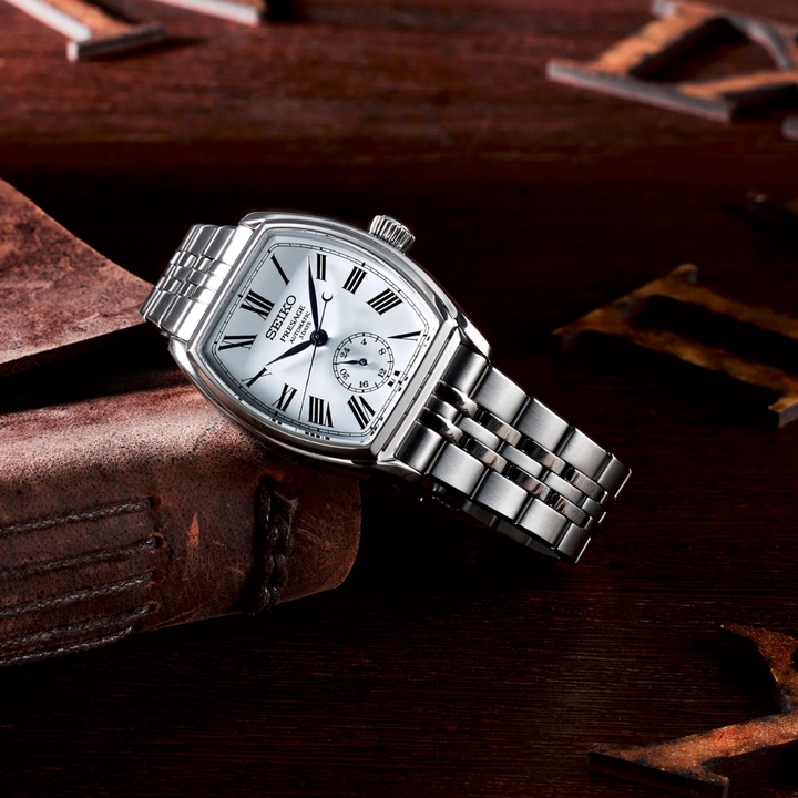 Seiko Presage SPB537J1 tonneau automatic watch on leather surface — white enamel dial with Roman numerals, 24-hour subdial, blue hands, silver bracelet, heritage Roman numeral backdrop