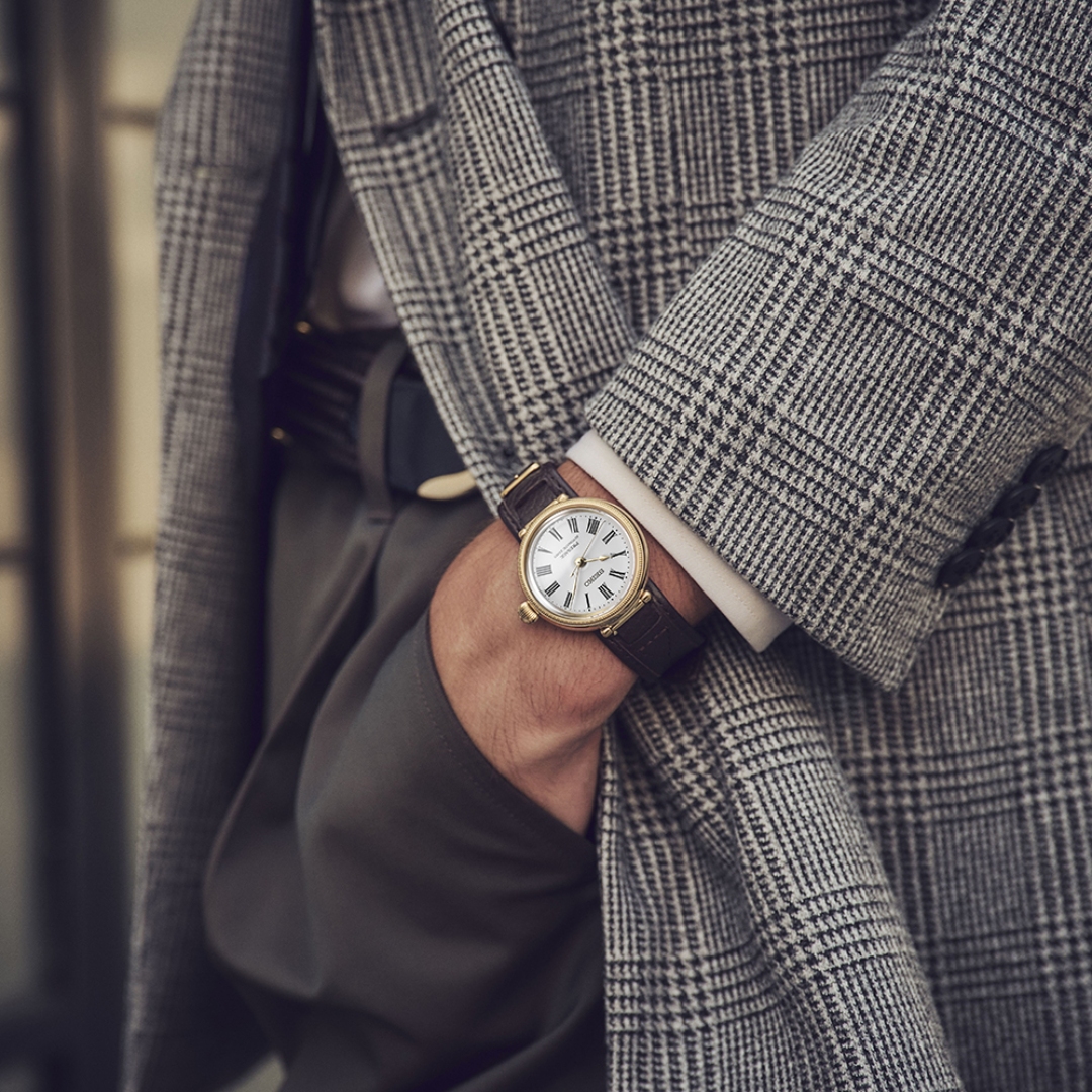 Seiko Presage SPB538J1 145th Anniversary automatic watch worn on wrist — white hand-enamelled dial with Roman numerals, gold-coloured millegrain case, dark brown leather strap, grey check jacket