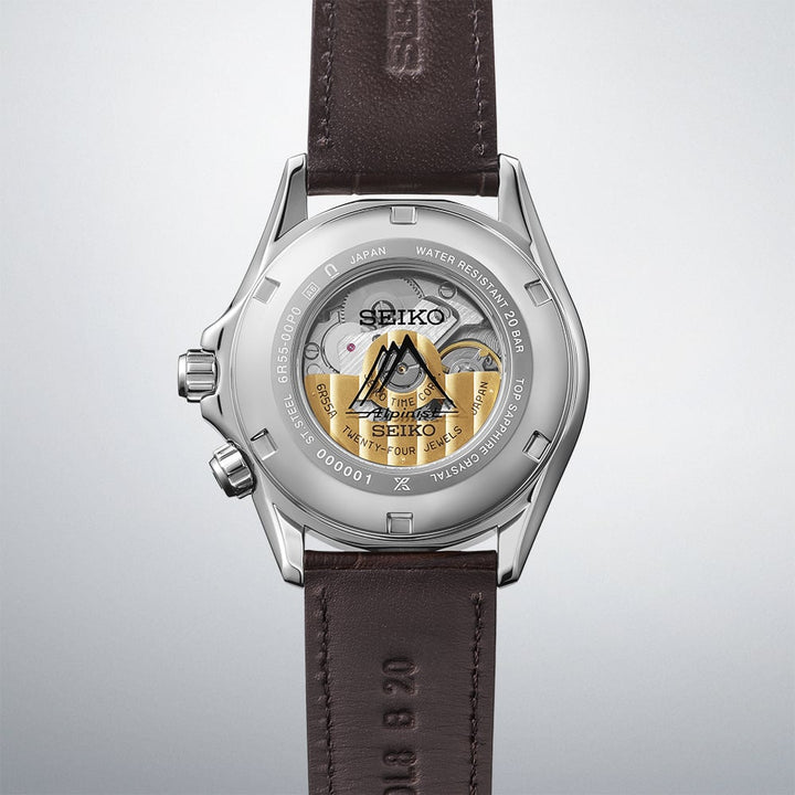 Seiko Prospex Alpinist SPB507J1 open caseback revealing the 6R55 automatic movement with gold Alpinist mountain rotor motif, serial number engraving, and dark brown cow leather strap on gradient grey background