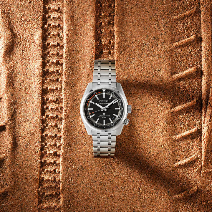 Seiko Prospex Speedtimer SPB515J1 automatic watch laid flat on sand terrain — black dial with orange countdown inner ring and stainless steel bracelet